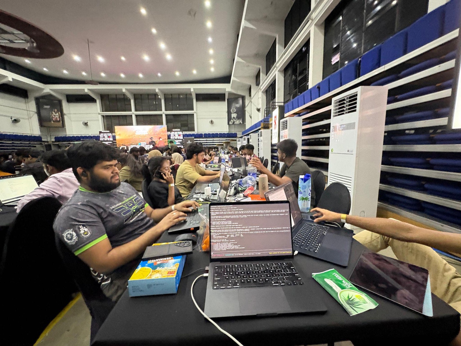 Coding at the BitByBit table with Gemini notes on screen and the stadium behind